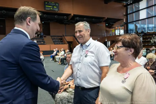 Spotkanie trzech osób, uściski dłoni i uśmiechy w tle sala z publicznością.