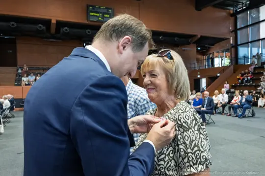 Mężczyzna przypina odznaczenie kobiecie podczas uroczystości w sali.