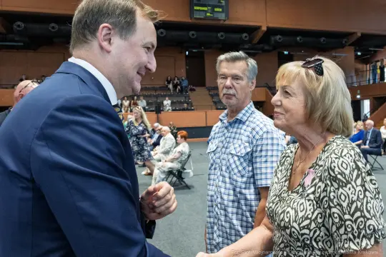Spotkanie dwóch osób z mężczyzną w garniturze, uściski dłoni i rozmowa. W tle publiczność.