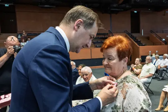 Mężczyzna przypina odznaczenie kobiecie podczas uroczystości. W tle siedzą inni uczestnicy.