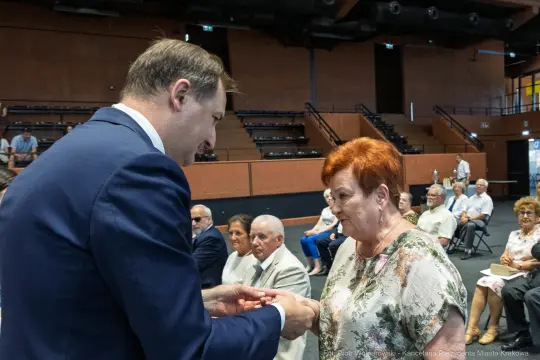 Wręczenie odznaczenia przez mężczyznę starszej kobiecie w sali z publicznością.