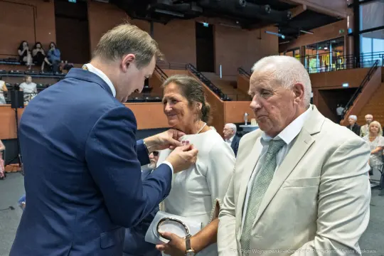 Uroczystość wręczenia odznaczenia kobiecie przez mężczyznę w eleganckim garniturze.
