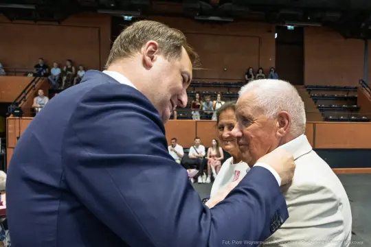 Uroczystość wręczenia odznaczeń, mężczyzna przypina medal starszemu mężczyźnie.