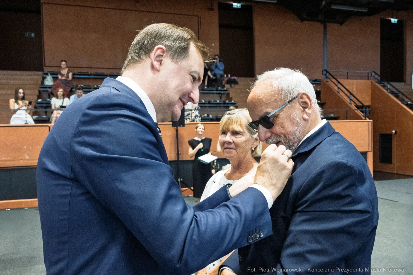 Mężczyzna przypina odznaczenie do marynarki innego mężczyzny, w tle kobieta obserwuje.  Autor: Piotr Wojnarowski