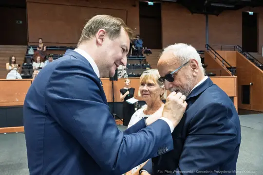Mężczyzna przypina odznaczenie do marynarki innego mężczyzny, w tle kobieta obserwuje.