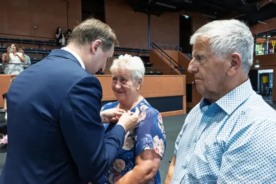 Wręczenie odznaczenia starszej parze przez mężczyznę w garniturze.