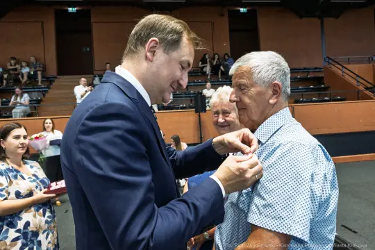 Uroczystość wręczenia odznaczeń, mężczyzna przypina medal starszemu mężczyźnie.