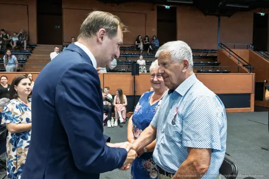 Mężczyzna w garniturze podaje rękę starszemu mężczyźnie, obok stoi kobieta. W tle widownia.