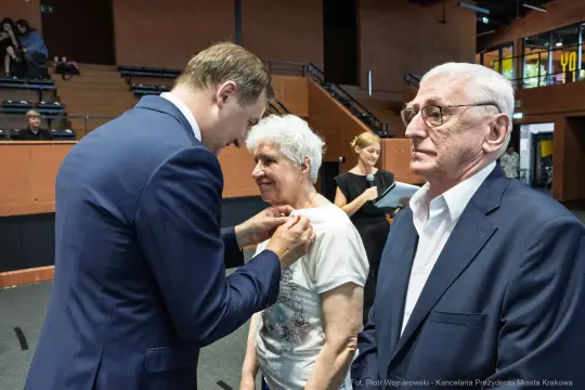 Wręczenie odznaczenia starszej kobiecie przez mężczyznę w garniturze, obok stoi starszy mężczyzna.