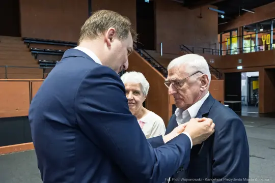 Mężczyzna przypina odznaczenie starszemu mężczyźnie, w tle kobieta obserwuje.