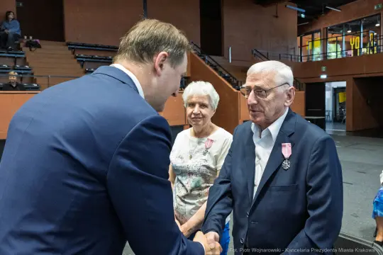 Uroczystość wręczenia odznaczeń, mężczyzna w garniturze podaje rękę starszemu mężczyźnie z medalem.