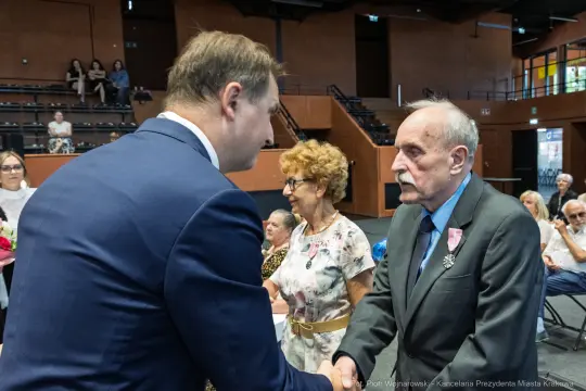 Uroczystość wręczenia odznaczeń, mężczyzna w garniturze podaje rękę starszemu mężczyźnie.