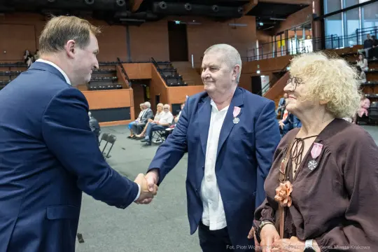 Uroczystość wręczenia odznaczeń, uściski dłoni, osoby w eleganckich strojach.