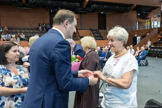 Wręczenie odznaczenia przez mężczyznę kobiecie podczas uroczystości w sali. W tle goście z bukietami kwiatów.