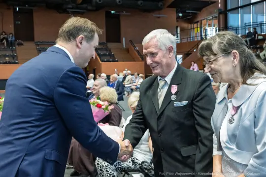Uroczystość wręczenia odznaczeń, mężczyzna w garniturze podaje rękę starszemu mężczyźnie.