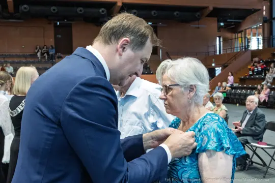 Wręczenie odznaczenia kobiecie przez mężczyznę w formalnym stroju.