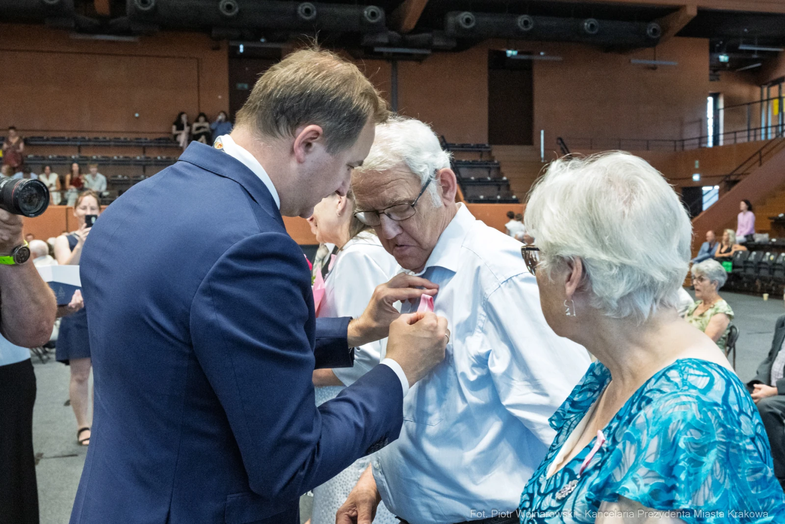 Mężczyzna przypina odznaczenie starszemu mężczyźnie, obok stoi kobieta.  Autor: Piotr Wojnarowski