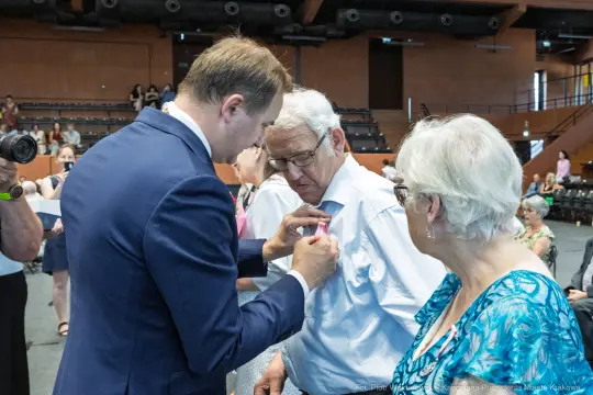 Mężczyzna przypina odznaczenie starszemu mężczyźnie, obok stoi kobieta.
