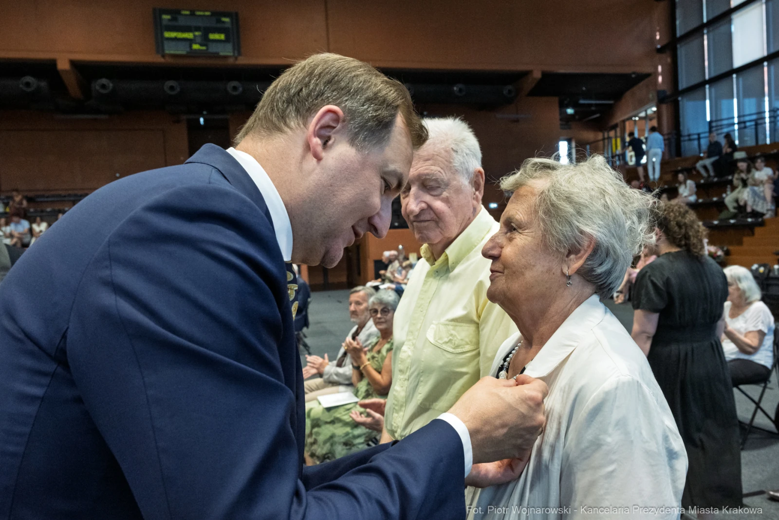 Mężczyzna wręcza odznaczenie starszej kobiecie w towarzystwie mężczyzny. W tle widoczna publiczność.  Autor: Piotr Wojnarowski