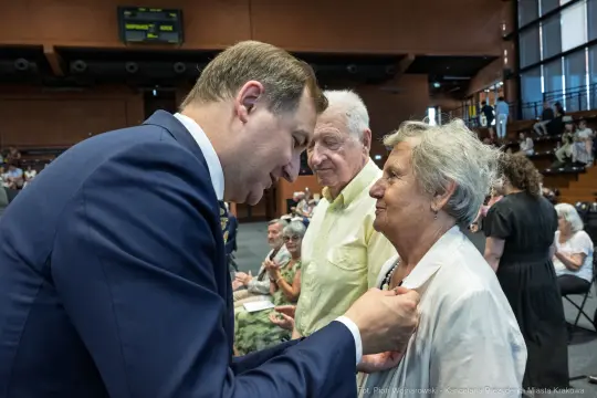 Mężczyzna wręcza odznaczenie starszej kobiecie w towarzystwie mężczyzny. W tle widoczna publiczność.