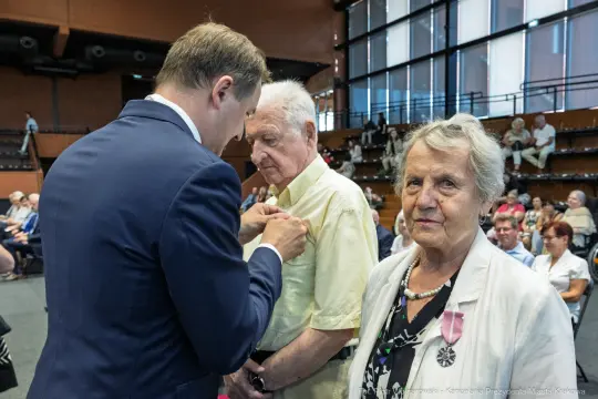 Uroczystość wręczenia odznaczeń, mężczyzna przypina medal starszemu mężczyźnie, obok stoi kobieta.