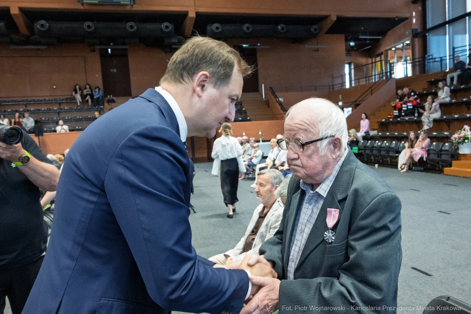 Mężczyzna w garniturze ściska dłoń starszemu mężczyźnie z odznaczeniem. W tle siedzą inni uczestnicy wydarzenia.  Autor: Piotr Wojnarowski