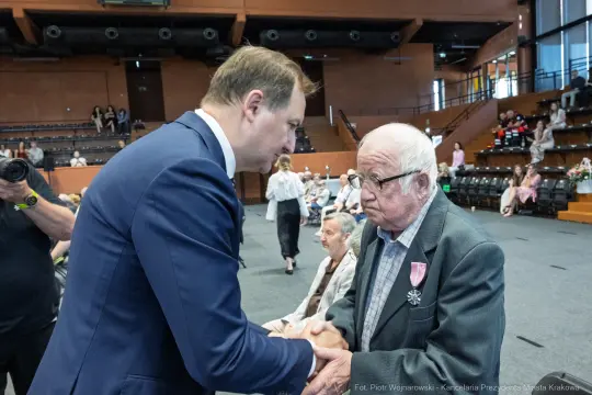 Mężczyzna w garniturze ściska dłoń starszemu mężczyźnie z odznaczeniem. W tle siedzą inni uczestnicy wydarzenia.