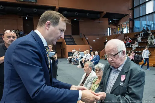 Wręczenie odznaczenia przez mężczyznę w garniturze starszemu mężczyźnie z medalem. W tle widownia.