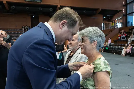 Wręczenie odznaczenia przez mężczyznę starszej kobiecie w towarzystwie innego mężczyzny.