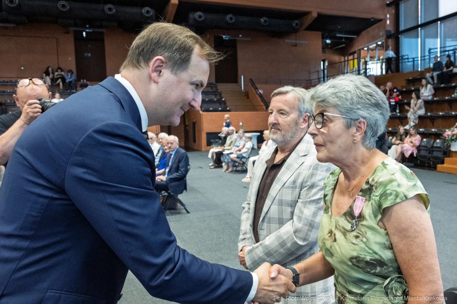 Mężczyzna w garniturze podaje rękę starszej kobiecie, w tle siedzą inni uczestnicy wydarzenia.  Autor: Piotr Wojnarowski