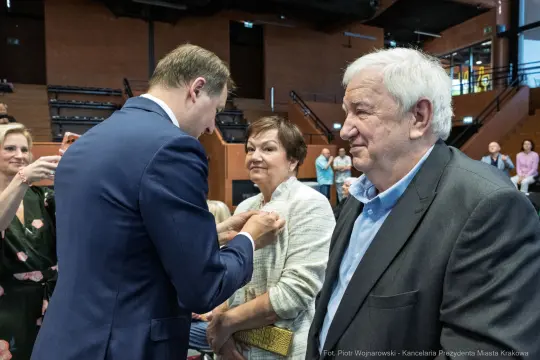 Wręczenie odznaczenia przez mężczyznę kobiecie i mężczyźnie w sali.