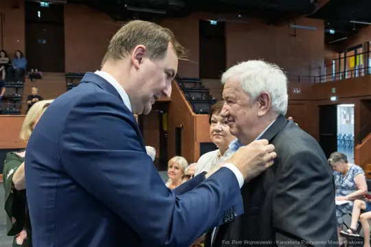Mężczyzna przypina odznaczenie innemu mężczyźnie w towarzystwie osób w tle.