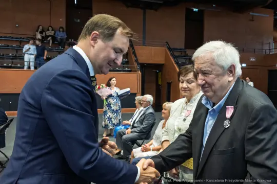 Uroczystość wręczenia odznaczeń, mężczyzna w garniturze podaje rękę starszemu mężczyźnie.
