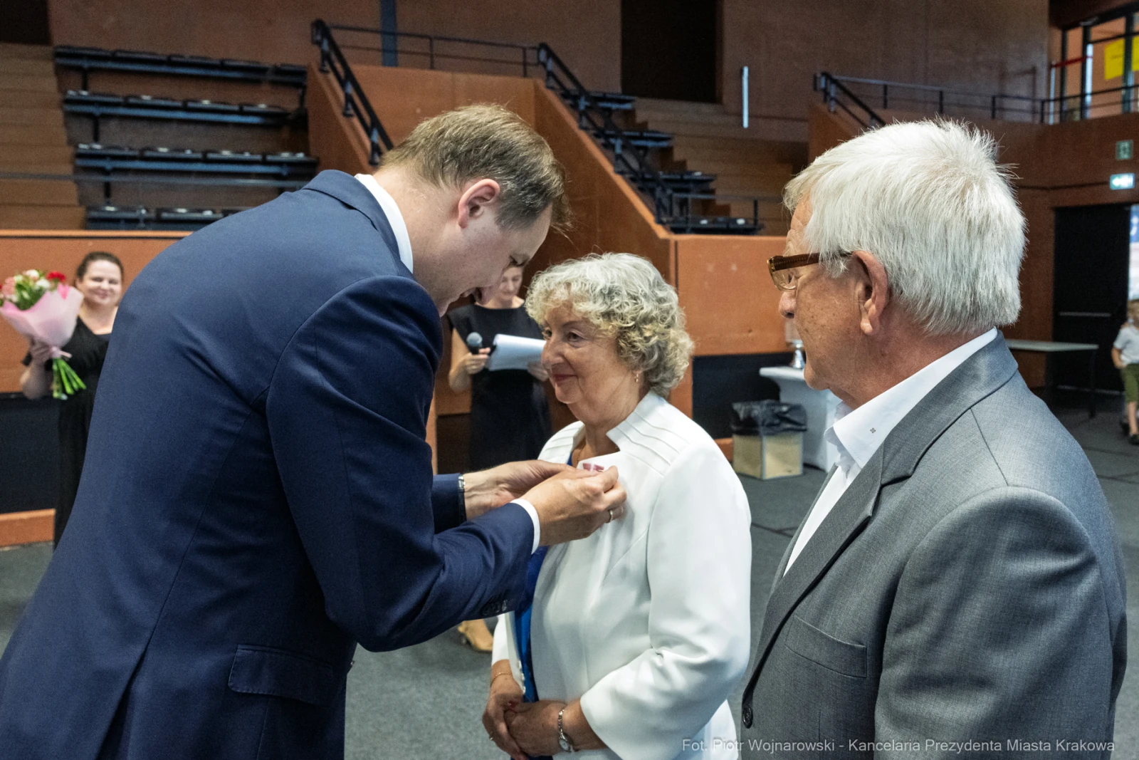 Uroczystość wręczenia odznaczenia, mężczyzna przypina medal kobiecie. Obok stoi mężczyzna.  Autor: Piotr Wojnarowski
