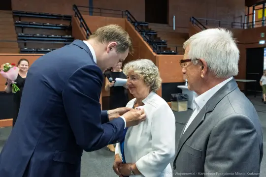 Uroczystość wręczenia odznaczenia, mężczyzna przypina medal kobiecie. Obok stoi mężczyzna.