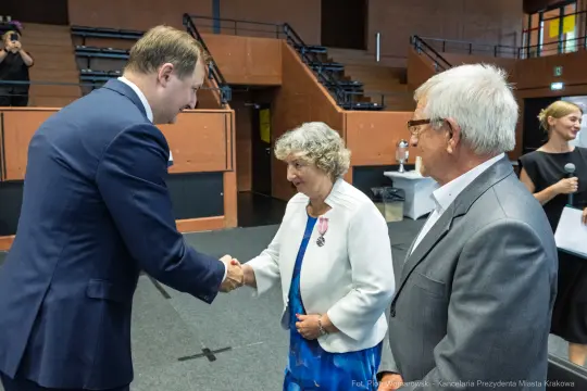 Uścisk dłoni między mężczyzną a kobietą, w tle inni uczestnicy wydarzenia.