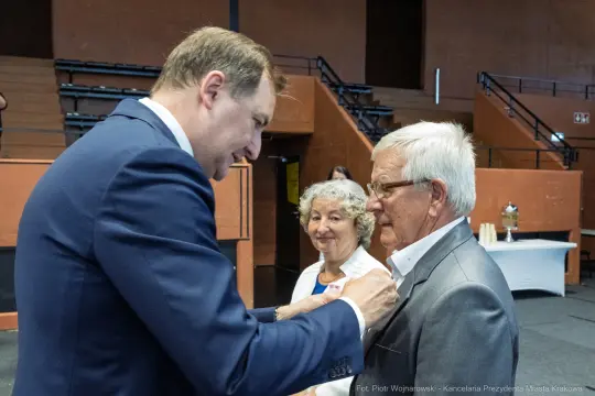 Uroczystość wręczenia odznaczeń, mężczyzna przypina medal starszemu mężczyźnie. W tle kobieta.