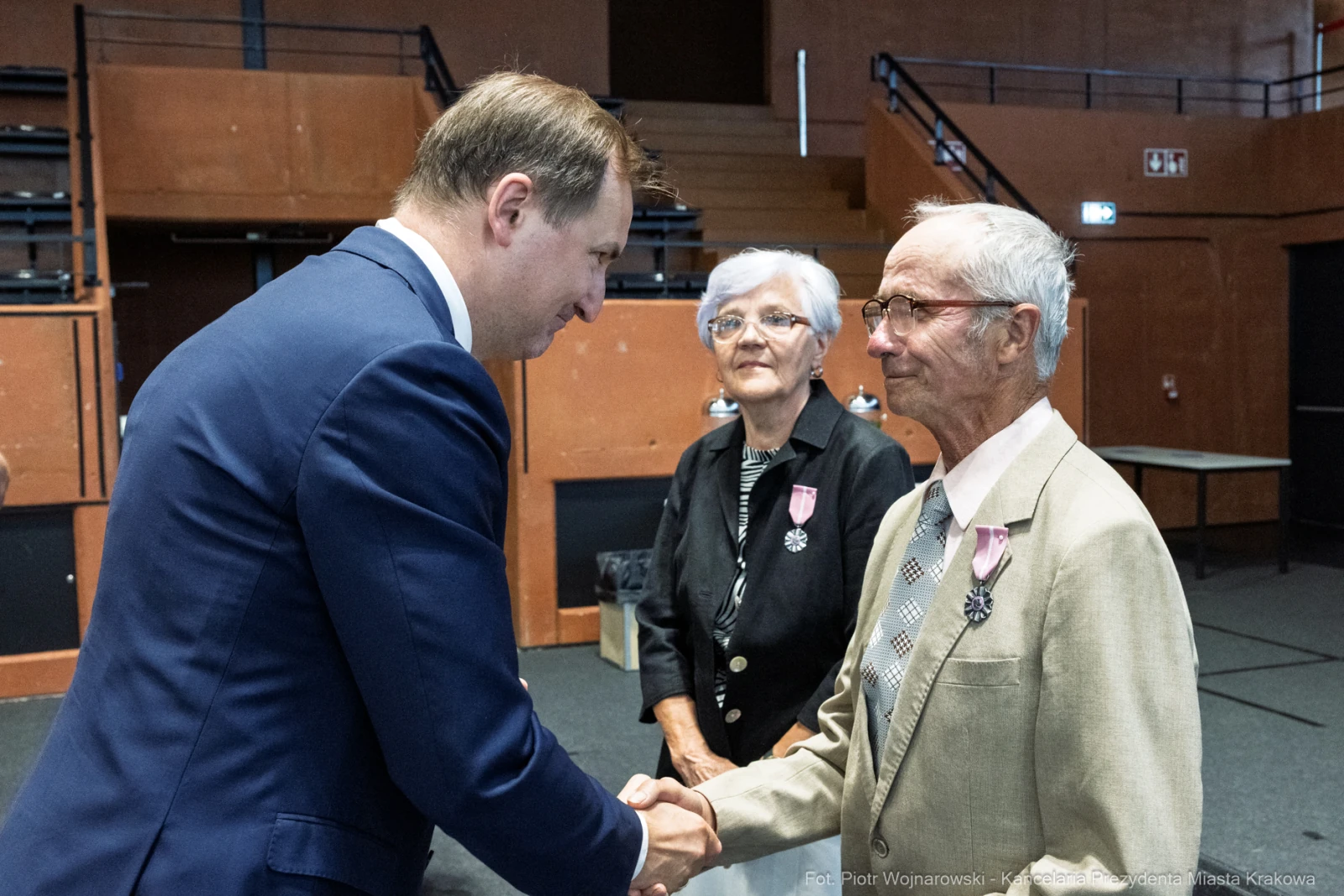 Uroczystość wręczenia odznaczeń, mężczyzna w garniturze wita starszego pana z medalem.  Autor: Piotr Wojnarowski