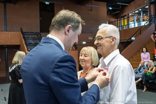Mężczyzna przypina odznaczenie starszemu mężczyźnie w obecności kobiety.