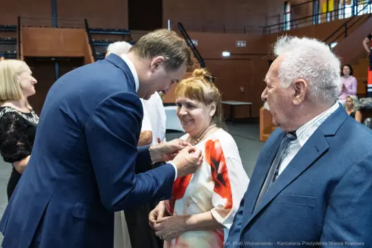 Wręczenie odznaczenia kobiecie przez mężczyznę w formalnym otoczeniu. Obecni także inni goście.