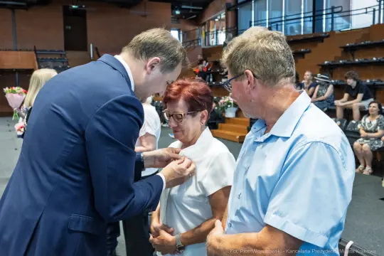 Wręczenie odznaczenia przez mężczyznę kobiecie w obecności innego mężczyzny.