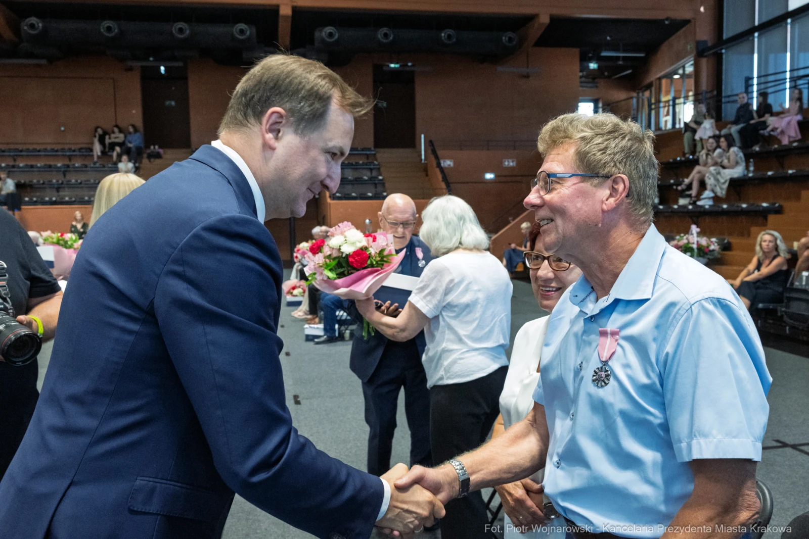 Mężczyzna w garniturze uścisnął dłoń starszemu mężczyźnie z odznaczeniem. W tle widoczni goście.  Autor: Piotr Wojnarowski