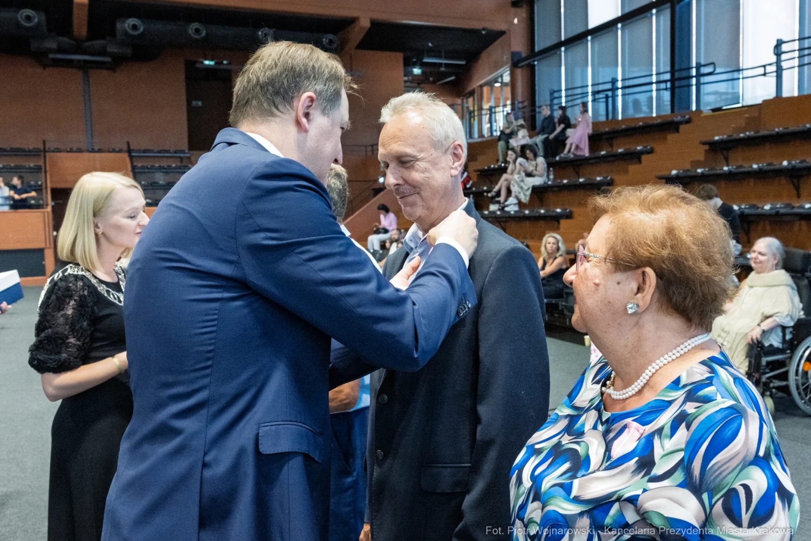 Mężczyzna przypina odznaczenie innemu mężczyźnie w obecności kobiety.  Autor: Piotr Wojnarowski