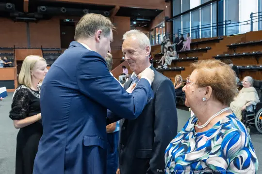 Mężczyzna przypina odznaczenie innemu mężczyźnie w obecności kobiety.