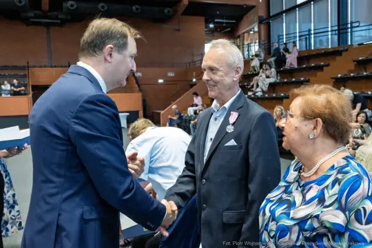 Uroczystość wręczenia odznaczeń, mężczyzna w garniturze wita odznaczonego i jego żonę.