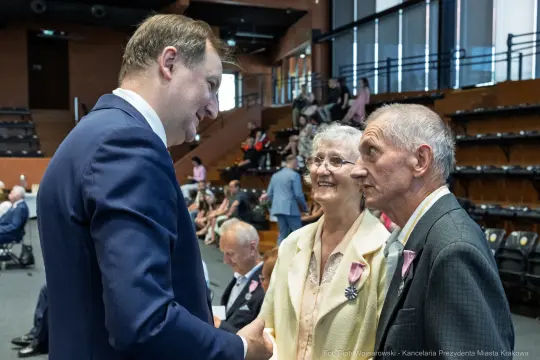 Spotkanie trzech osób w formalnych strojach, rozmowa w sali z widownią w tle.