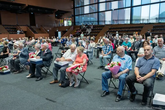 Grupa ludzi siedzących na krzesłach w sali, część z kwiatami w rękach.