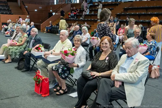 Grupa osób siedzących na krzesłach, trzymających kwiaty i prezenty, w tle widoczne inne osoby.