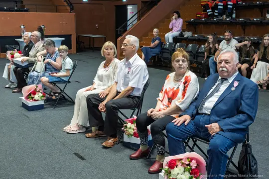Grupa osób siedzących na krzesłach, w tle widoczni widzowie. Uczestnicy z kwiatami w rękach.