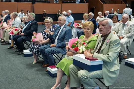 Grupa osób siedzących na uroczystości, trzymających kwiaty i upominki.